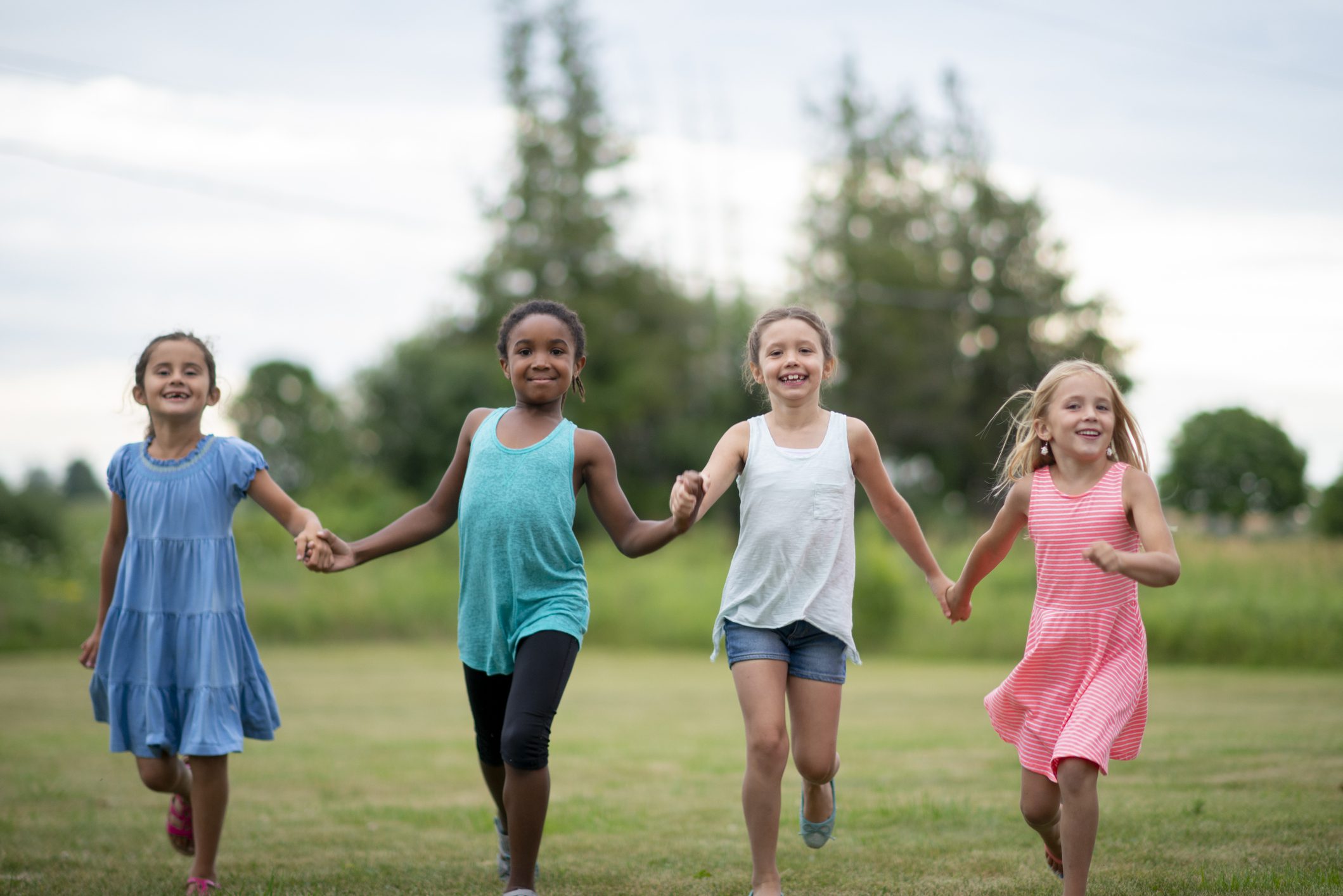 A small group of four school aged children, hold hands as they run through the grass together on a sunny summer day.