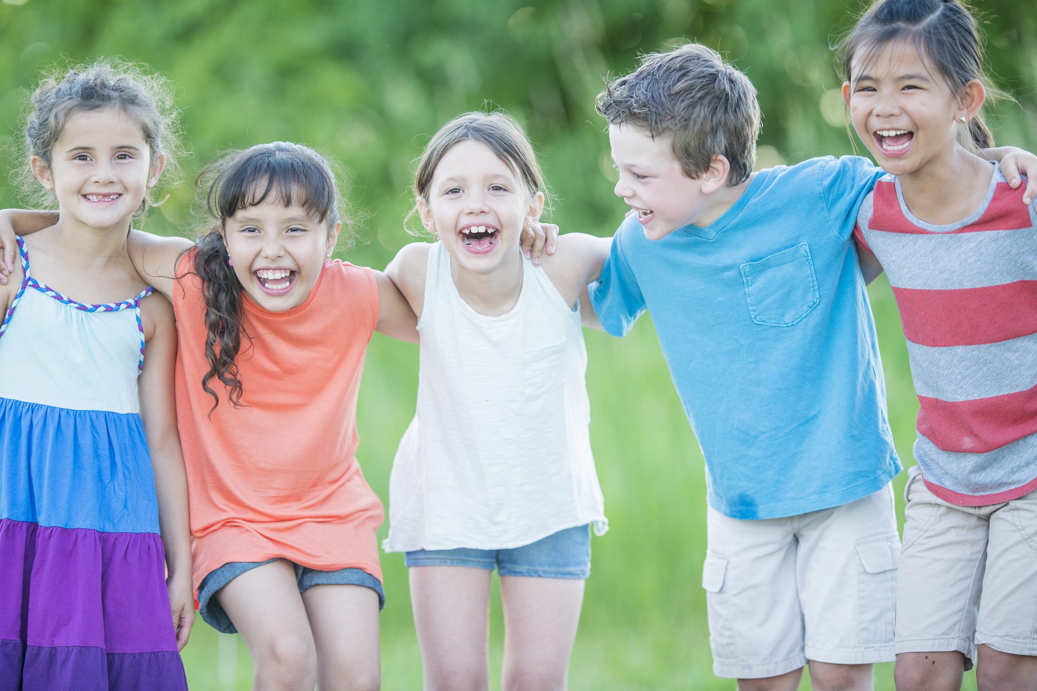 A multi-ethnic group of elementary school children are outside on a sunny day.  They are wearing casual clothing. They are smiling and laughing while posing together.